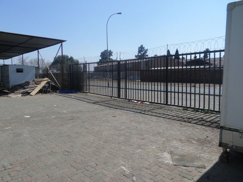 Industrial yard area featuring a large metal security gate with razor wire, paved ground, and a covered storage structure.