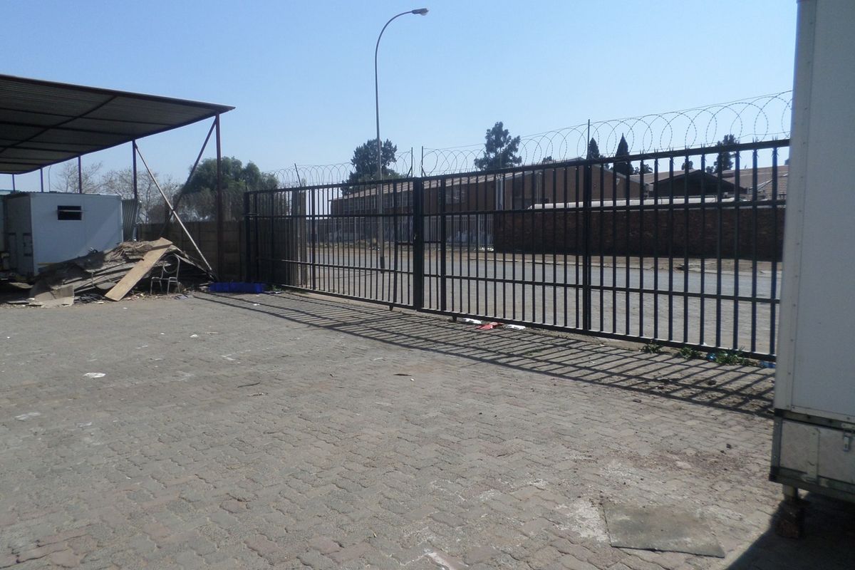 Industrial yard area featuring a large metal security gate with razor wire, paved ground, and a covered storage structure.