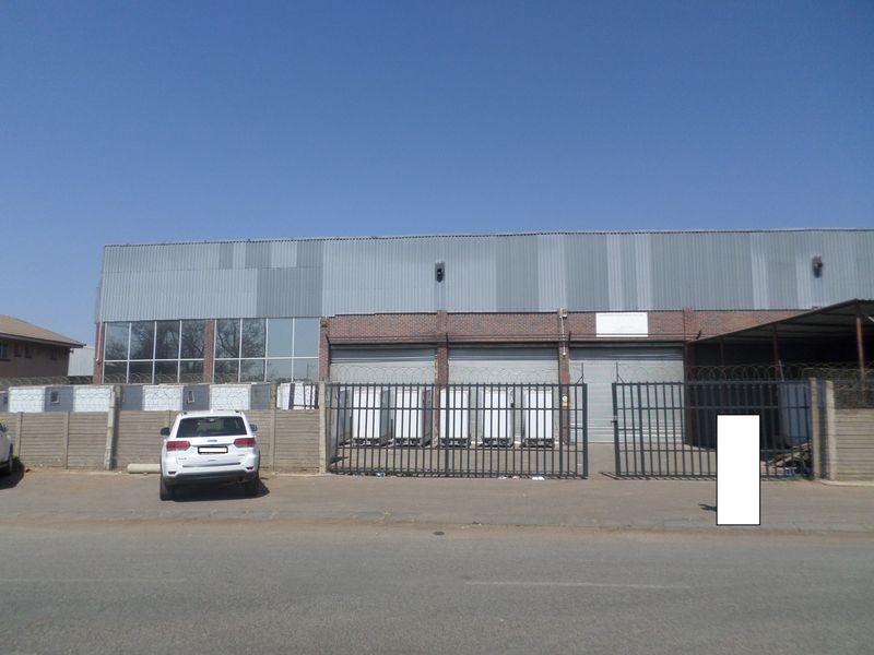 Front elevation of an industrial warehouse facility featuring corrugated metal cladding, brickwork, multiple roller shutter doors, and secure perimeter fencing.