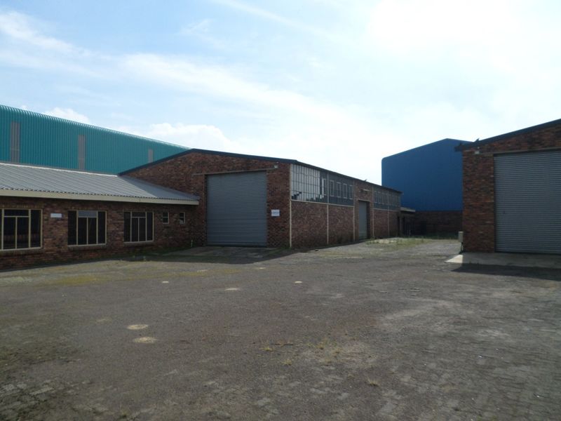 Exterior view of an industrial property featuring multiple warehouse buildings with roller shutter doors, brick facades, and a large paved yard area.