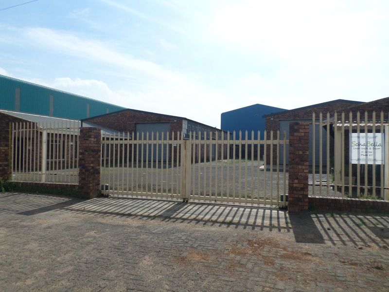 Front view of an industrial property featuring a secure gated entrance, paved driveway, and multiple warehouse units with roller shutter doors.