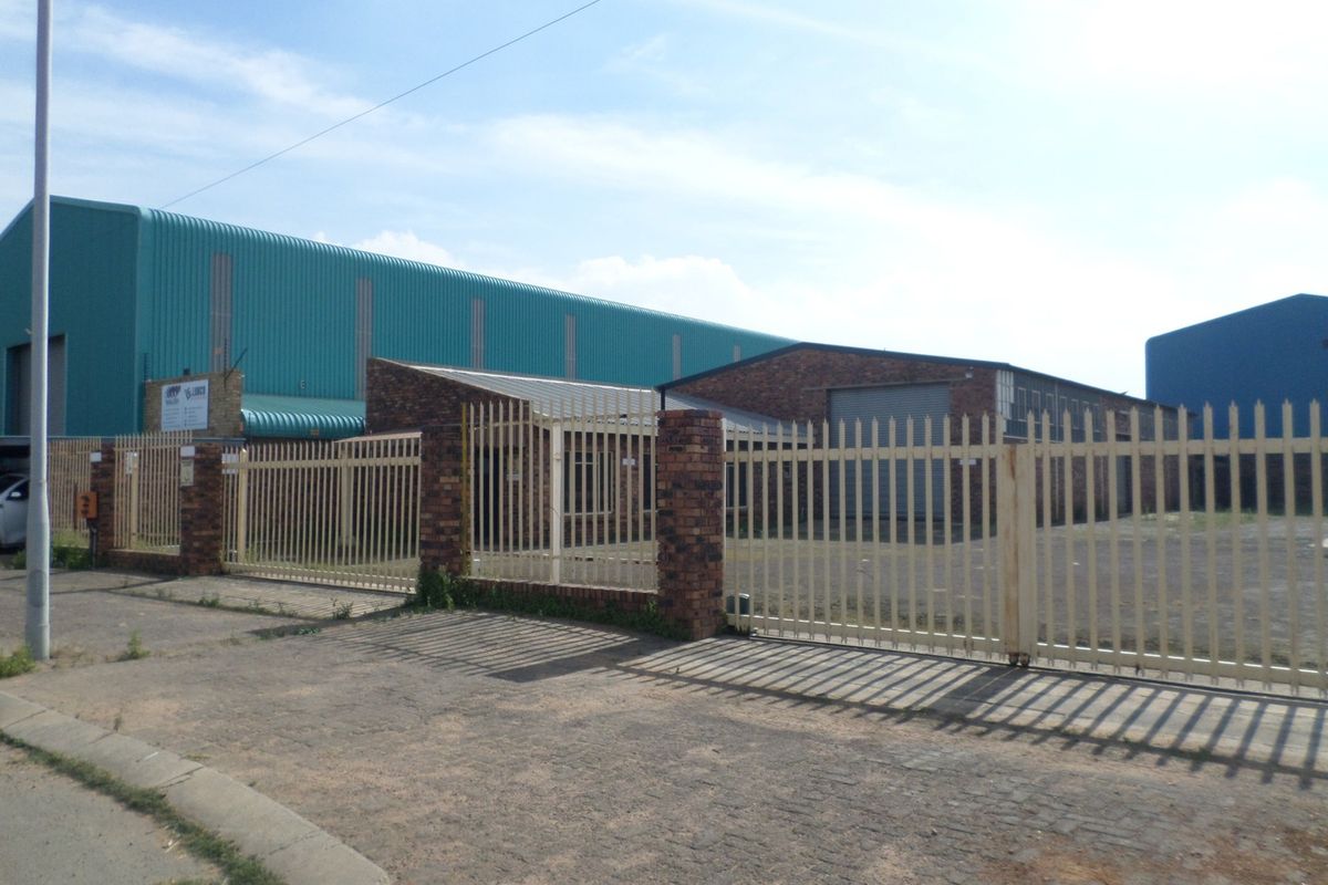 Industrial property exterior featuring a secure metal palisade fence, brick pillars, and a large warehouse structure with corrugated metal roofing and roller shutter access.