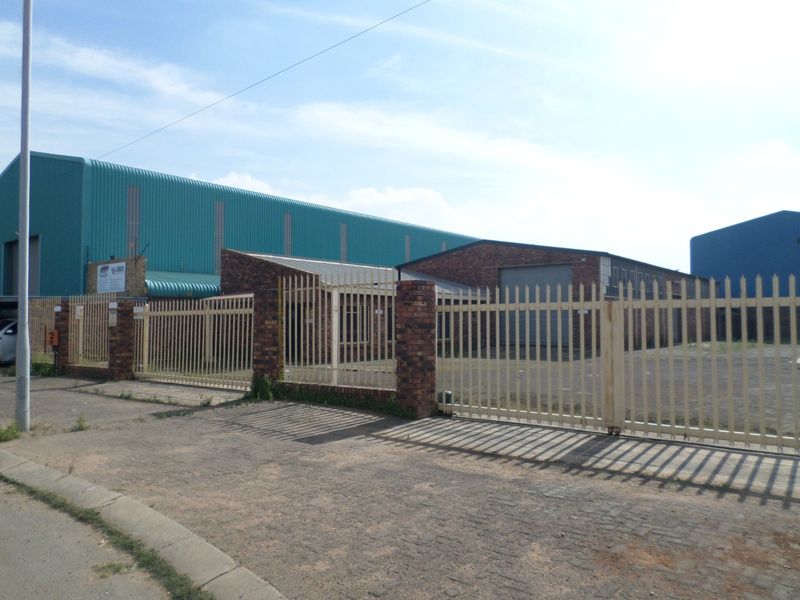 Industrial property exterior featuring a secure metal palisade fence, brick pillars, and a large warehouse structure with corrugated metal roofing and roller shutter access.