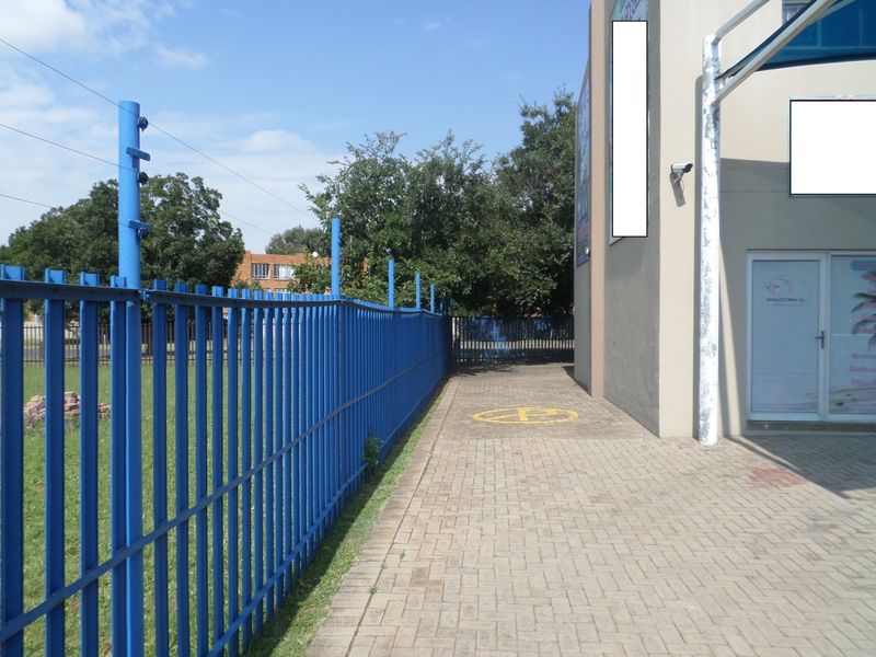 Exterior side view of the commercial property featuring a paved walkway, secure metal perimeter fencing with electric wires, and building-mounted security surveillance.