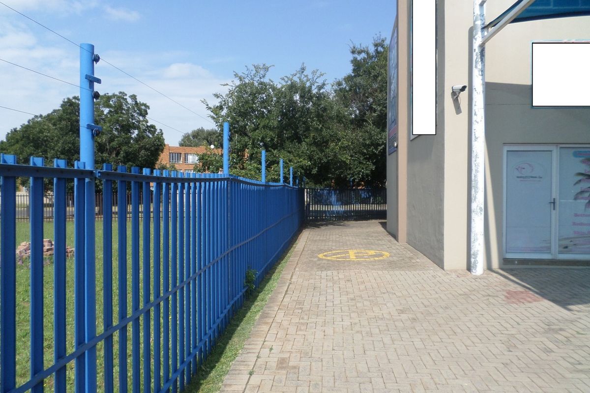Exterior side view of the commercial property featuring a paved walkway, secure metal perimeter fencing with electric wires, and building-mounted security surveillance.