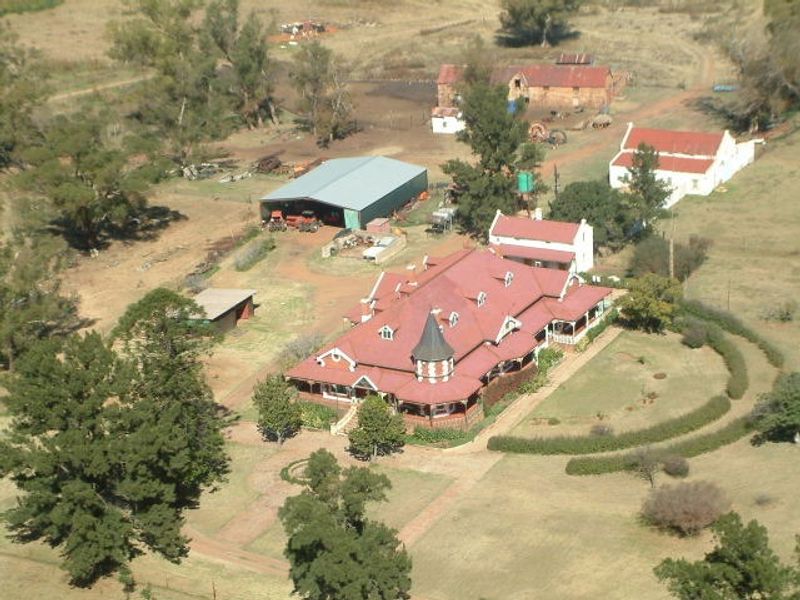 Aerial view of a large guest house property featuring a distinctive turret, expansive metal roofing, surrounding verandas, and multiple outbuildings set within a rural landscape.