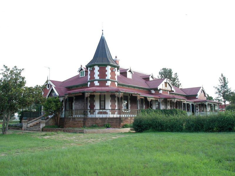 A historic guest house featuring a prominent turret, wrap-around veranda, and classic brick architecture set within a well-maintained garden.