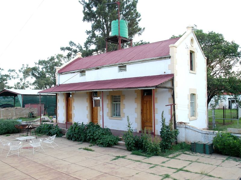 A rear view of a guest house featuring a corrugated metal roof, a covered veranda, and a paved patio area with an elevated water tank.