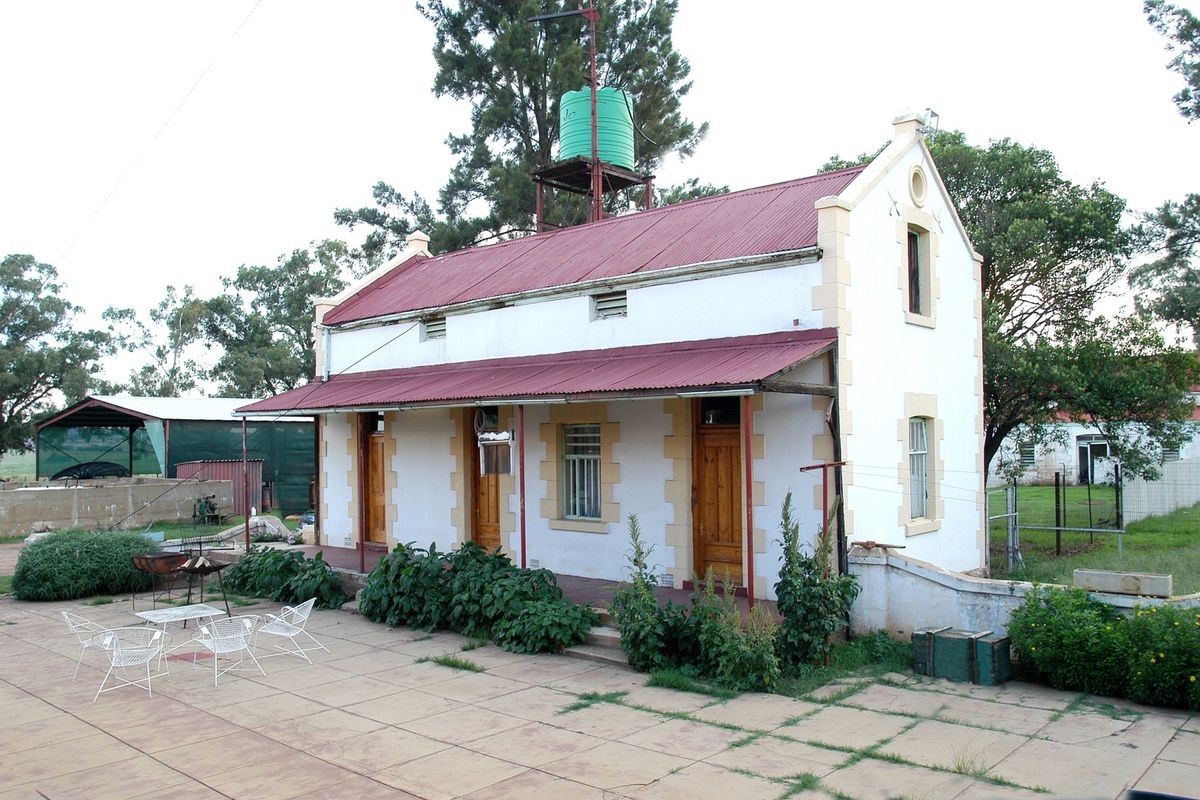 A rear view of a guest house featuring a corrugated metal roof, a covered veranda, and a paved patio area with an elevated water tank.
