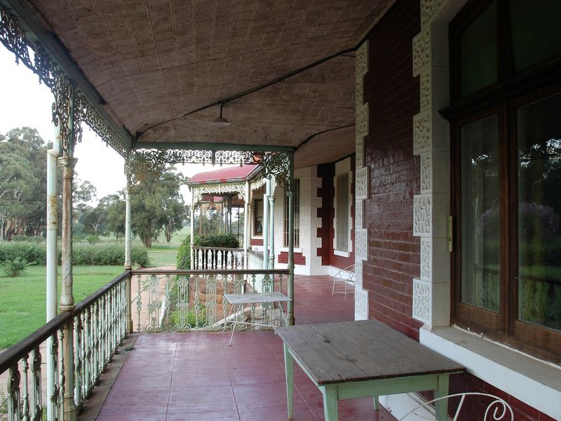 A covered wrap-around veranda featuring historic wrought iron detailing, tiled flooring, and views over the surrounding garden grounds.