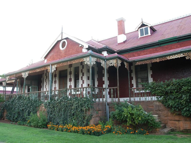 A historic brick guest house featuring a classic veranda with ornate wrought iron fretwork, a corrugated iron roof, and established garden surroundings.