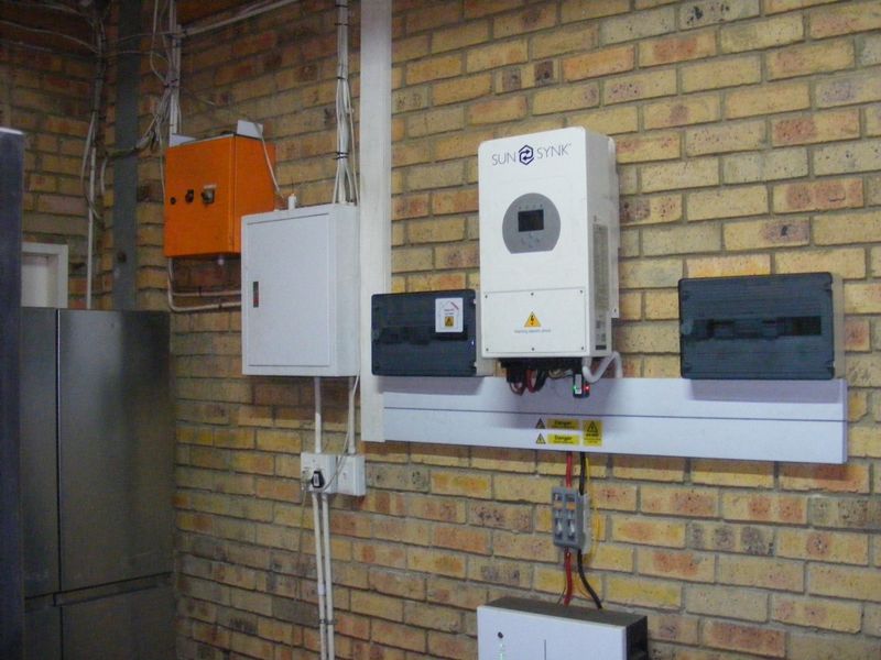 Utility area featuring a wall-mounted Sunsynk solar inverter and electrical distribution boards installed on an exposed brick wall.
