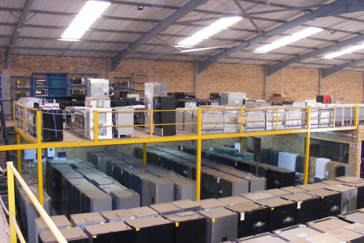 Large industrial warehouse space featuring a mezzanine level with steel safety railings, exposed brick walls, and high-clearance roof trusses with integrated skylights.