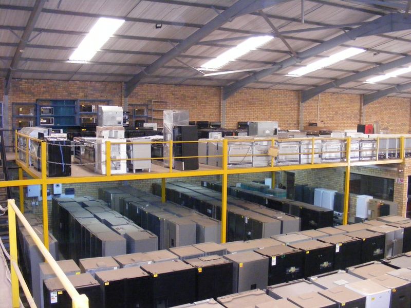 Large industrial warehouse space featuring a mezzanine level with steel safety railings, exposed brick walls, and high-clearance roof trusses with integrated skylights.