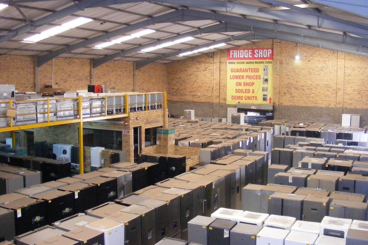 Large industrial warehouse space featuring exposed steel roof trusses, skylights, and a mezzanine level, currently utilized for appliance storage and retail.
