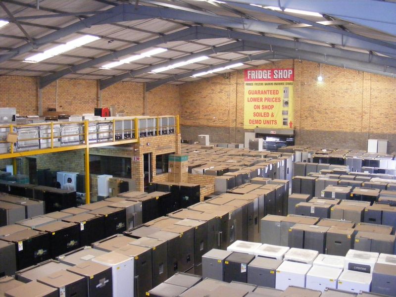 Large industrial warehouse space featuring exposed steel roof trusses, skylights, and a mezzanine level, currently utilized for appliance storage and retail.