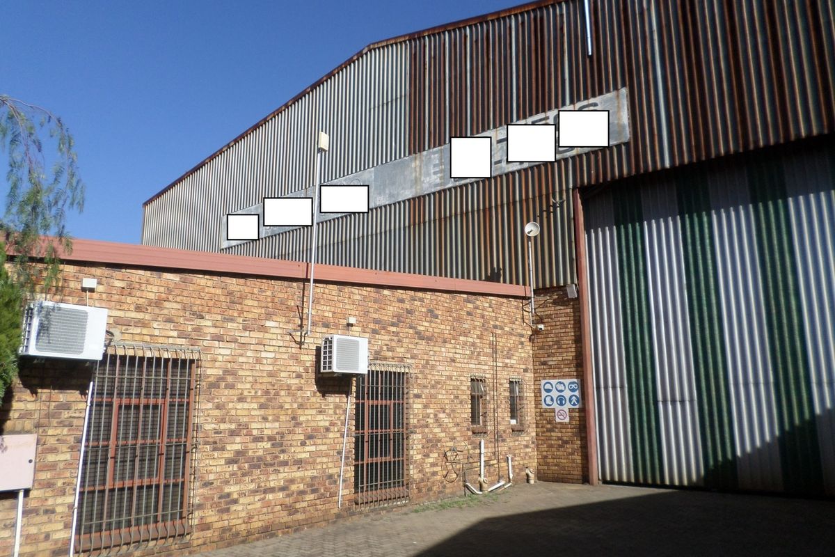 Rear industrial facade featuring face brick construction, corrugated metal cladding with visible corrosion, security-barred windows, and a large industrial roller shutter door.