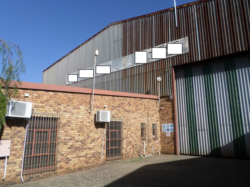 Rear industrial facade featuring face brick construction, corrugated metal cladding with visible corrosion, security-barred windows, and a large industrial roller shutter door.