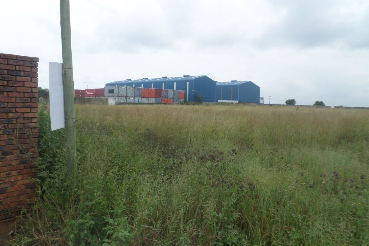 View of an industrial warehouse facility set behind an expansive, undeveloped grass field with perimeter brick walling.
