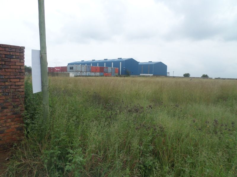 View of an industrial warehouse facility set behind an expansive, undeveloped grass field with perimeter brick walling.