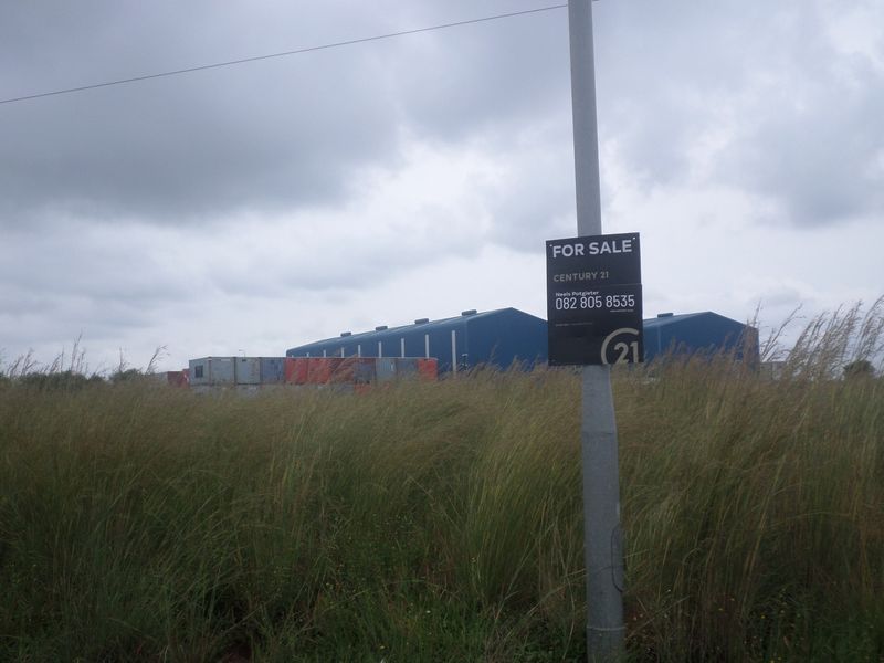 View of an industrial warehouse facility with metal cladding, situated on a large plot with tall grass and a for sale sign.