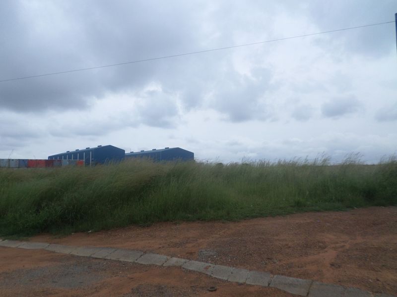 View of an industrial warehouse facility from an unpaved access road, featuring open land and overgrown grass.