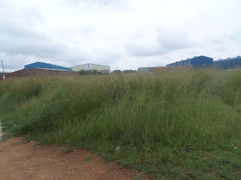 A vacant industrial plot featuring overgrown grass and a partial perimeter brick wall, situated within an established industrial park setting.