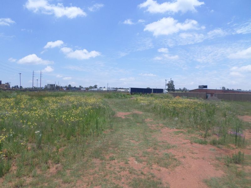 A view of a vacant commercial land plot featuring natural vegetation, perimeter fencing, and an unpaved access path under a clear blue sky.