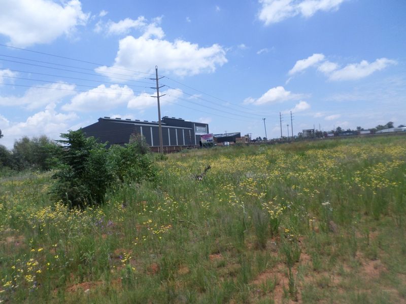 A view of a vacant commercial land plot with natural vegetation, featuring overhead power lines and a distant commercial warehouse structure.