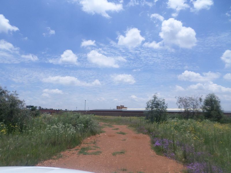 A view of a vacant commercial land plot featuring a dirt access path, natural vegetation, and distant perimeter fencing.