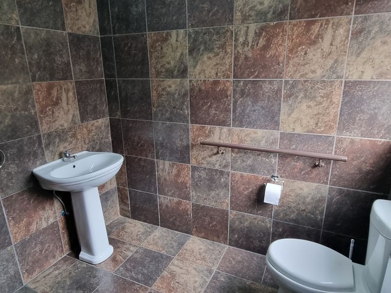 A functional bathroom featuring full-height wall tiling, a pedestal washbasin, a toilet, and a wall-mounted grab rail for accessibility.