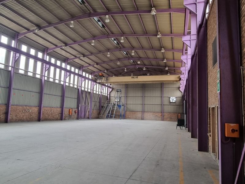 Spacious industrial warehouse featuring high ceilings, exposed steel trusses, an overhead crane, concrete flooring, and brick masonry walls.