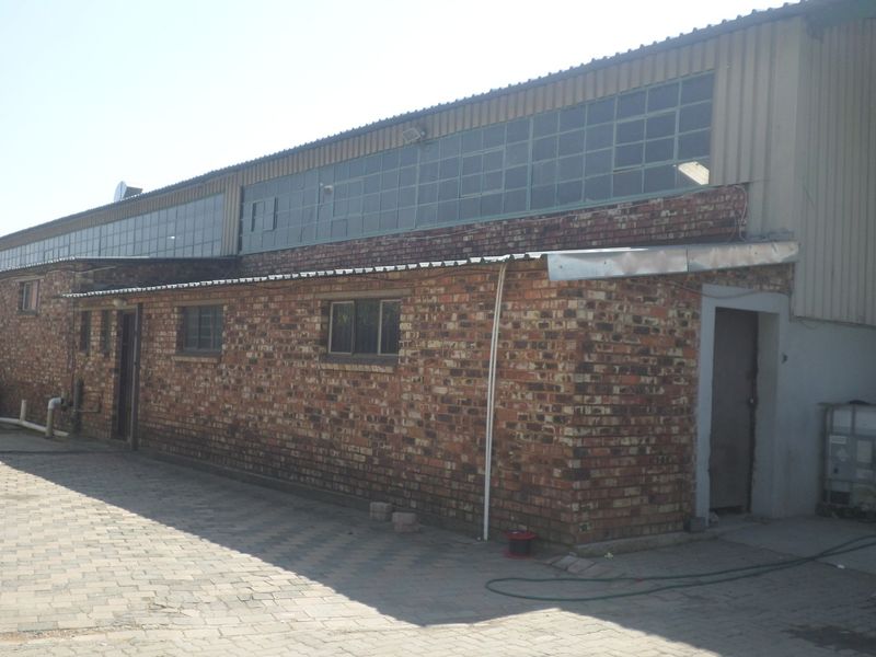 Rear exterior view of an industrial property featuring exposed brick walls, corrugated metal roofing, and large steel-framed windows overlooking a paved yard area.