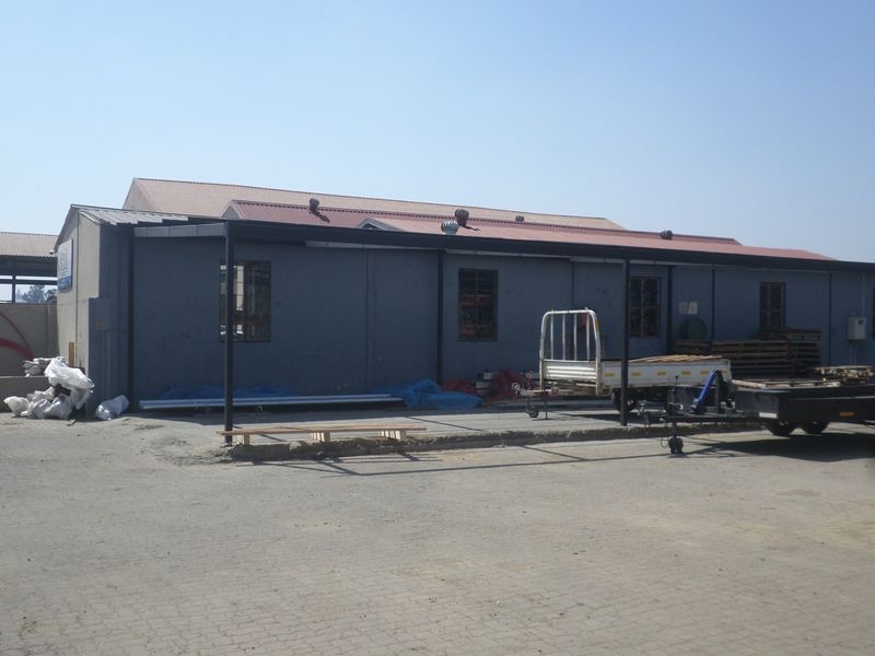 Rear view of an industrial warehouse building featuring a metal roof, roof ventilation turbines, and a covered outdoor area with paved yard space.