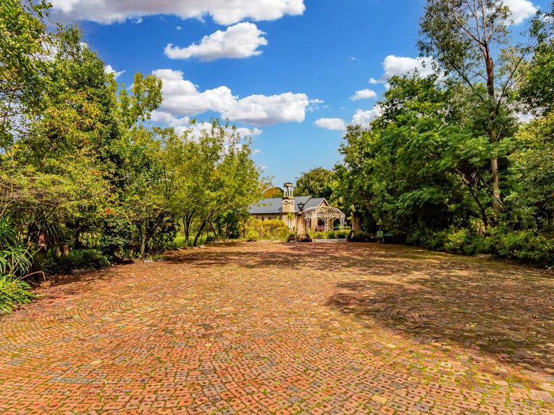 A wide, brick-paved driveway leading towards a stone-clad commercial building, surrounded by lush, mature trees and landscaped gardens.