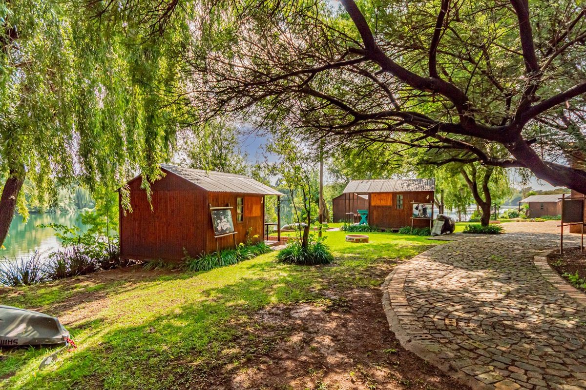 A scenic outdoor area featuring timber cabins situated along a waterfront, complemented by mature trees and a charming cobblestone pathway.