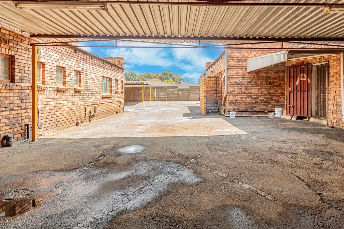 A paved industrial rear courtyard area featuring face brick walls, a covered parking section with a corrugated metal roof, and a secure steel gate.