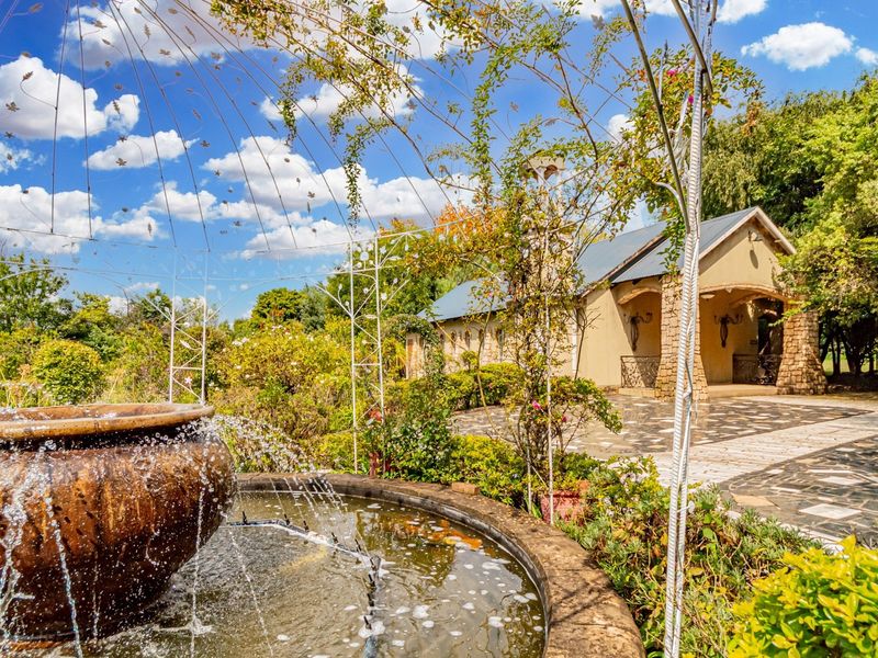 A beautifully landscaped garden featuring a stone fountain, a decorative metal gazebo frame, and a paved courtyard leading to a stone-accented commercial building.