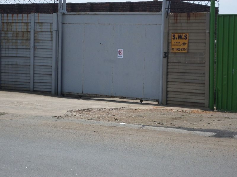 Industrial property entrance featuring a sliding gate, perimeter precast concrete walling with razor wire, and adjacent corrugated metal fencing.