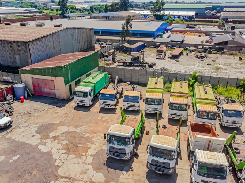 Aerial view of an industrial property featuring a large concrete yard, perimeter walling, and warehouse structures, currently utilized for heavy vehicle parking.