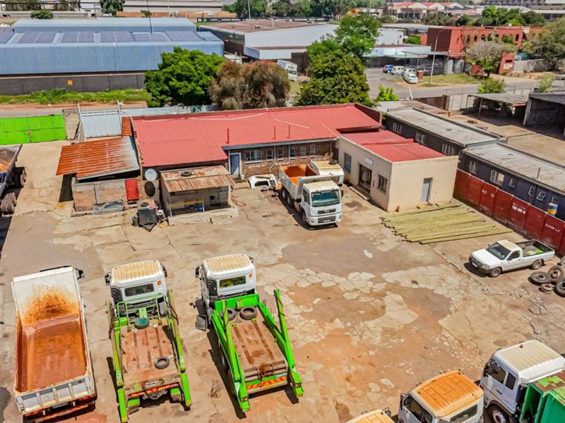 Aerial view of an industrial property featuring a large concrete yard, multiple warehouse structures with metal roofing, and perimeter security fencing.