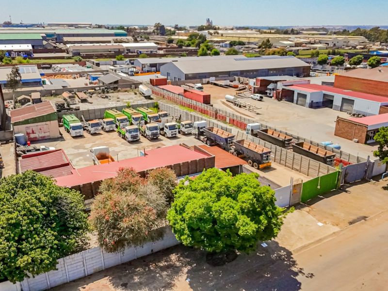 Aerial view of an industrial property featuring a large paved yard, perimeter walling, and multiple warehouse structures.