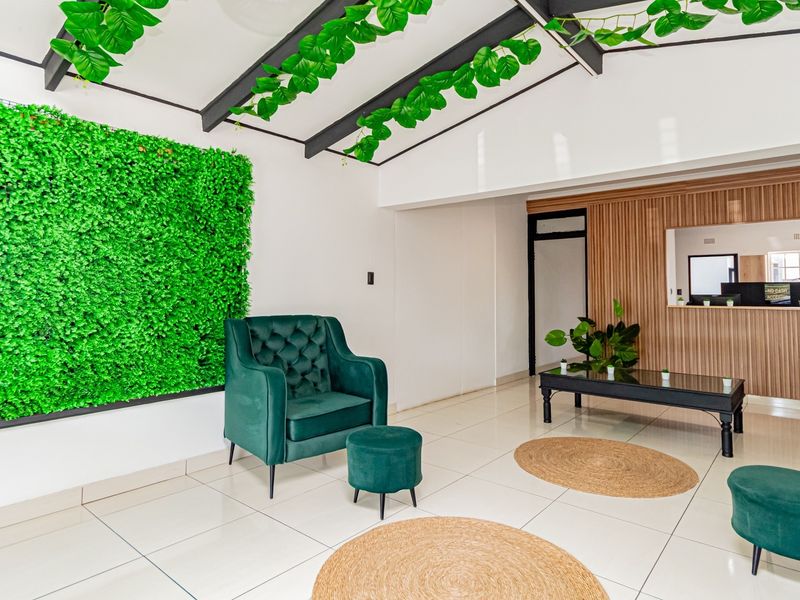 A modern, bright living area featuring tiled floors, exposed ceiling beams, and a decorative green wall, with a reception-style service window.