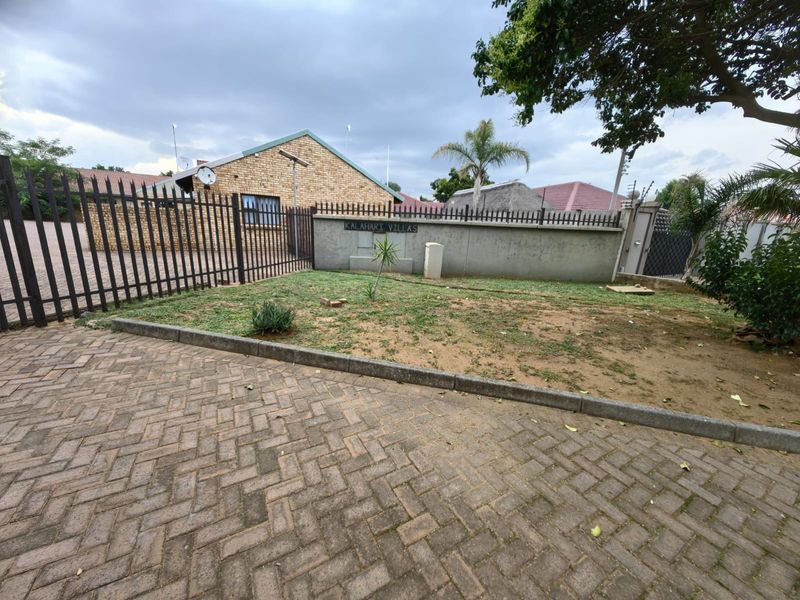 Front exterior view of the townhouse complex entrance featuring brick paving, metal security fencing, and a concrete boundary wall with electric fencing.