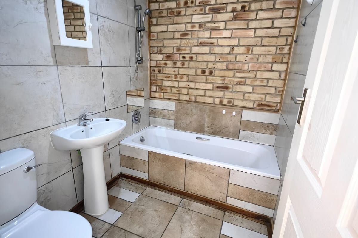 A functional bathroom featuring a pedestal basin, bathtub with shower attachment, and a distinctive exposed brick feature wall with neutral floor tiling.