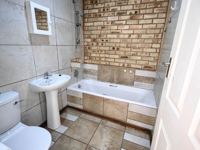 A functional bathroom featuring a pedestal basin, bathtub with shower attachment, and a distinctive exposed brick feature wall with neutral floor tiling.