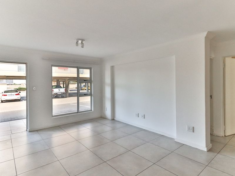 A bright, empty living room featuring neutral tiled flooring, clean white walls, and natural light from a large window and glass door.