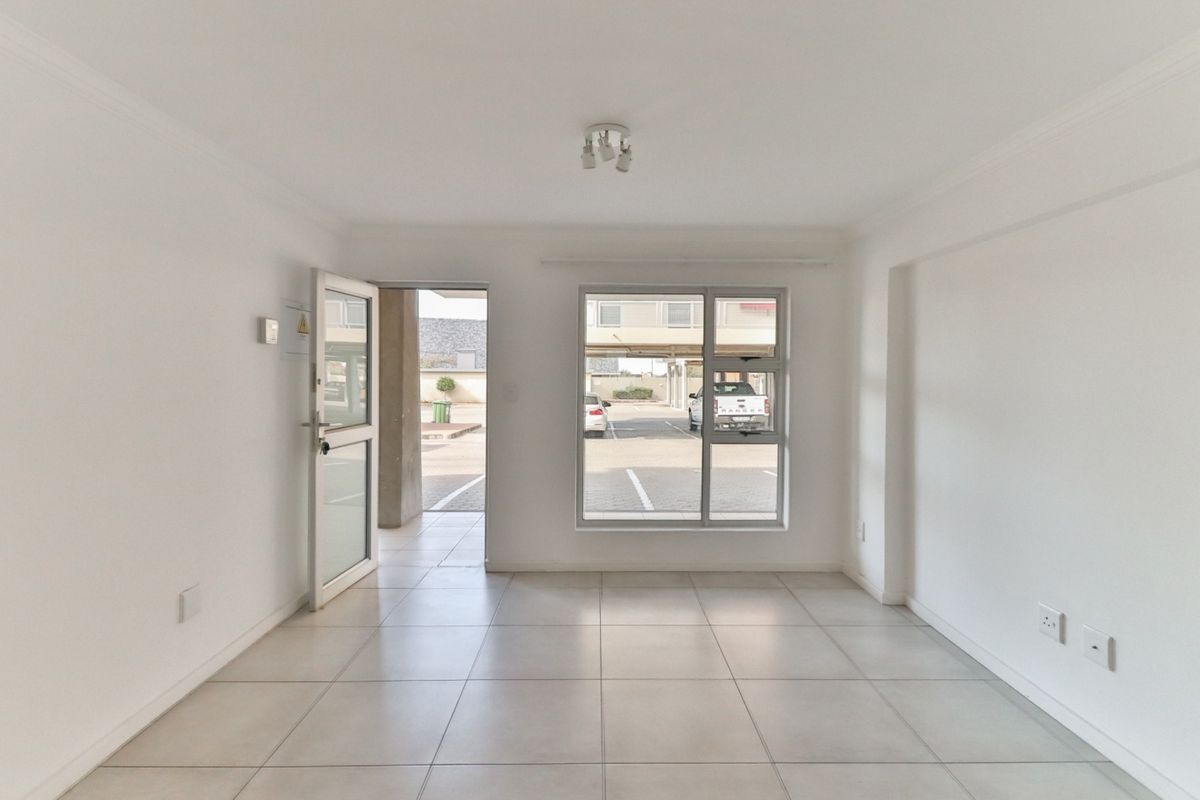 A bright, empty living area featuring neutral tiled flooring, clean white walls, and large aluminium-framed windows providing natural light and access to the exterior.