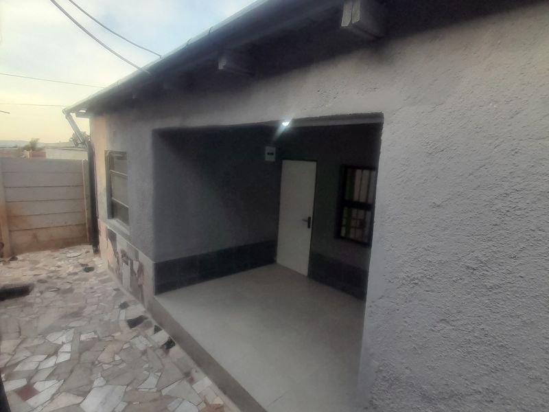 A covered patio area featuring tiled flooring and plastered walls, leading to a paved rear courtyard with security-fitted windows.