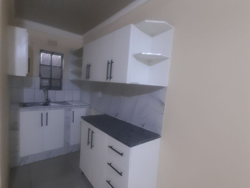 A clean, modern kitchen featuring white built-in cabinetry, dark countertops, tiled backsplash, and neutral floor tiles in a compact, functional layout.